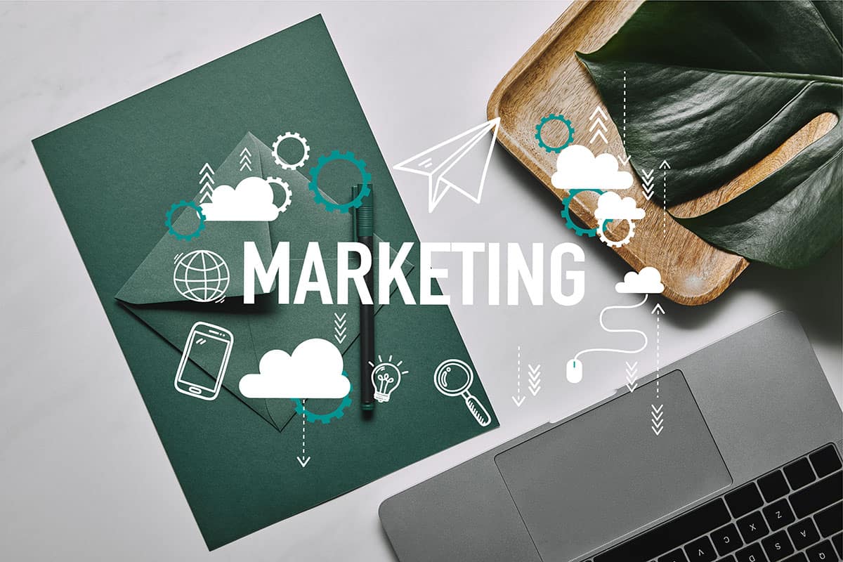 Marketing Concept A flat lay of a work desk featuring green stationery, a clipboard, a laptop, and a wooden tray with a leaf, overlaid with various marketing icons and the word "MARKETING" in bold white letters.